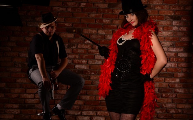 Woman in cabaret performers attire and man in mafioso attire with brick wall background